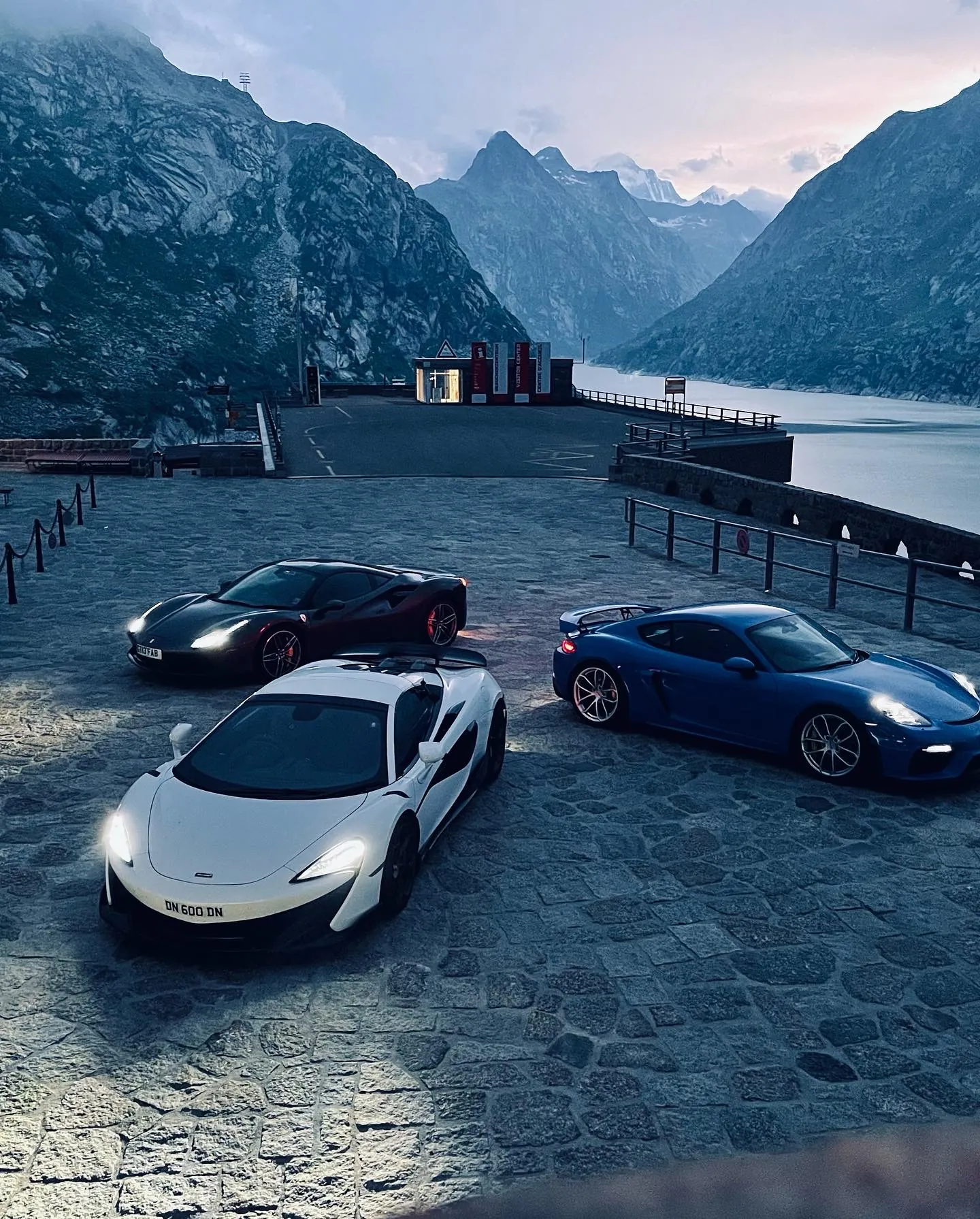 A Ferrari, a McLaren and a Porsche parked on the cobbles beside a mountain reservoir at dusk.