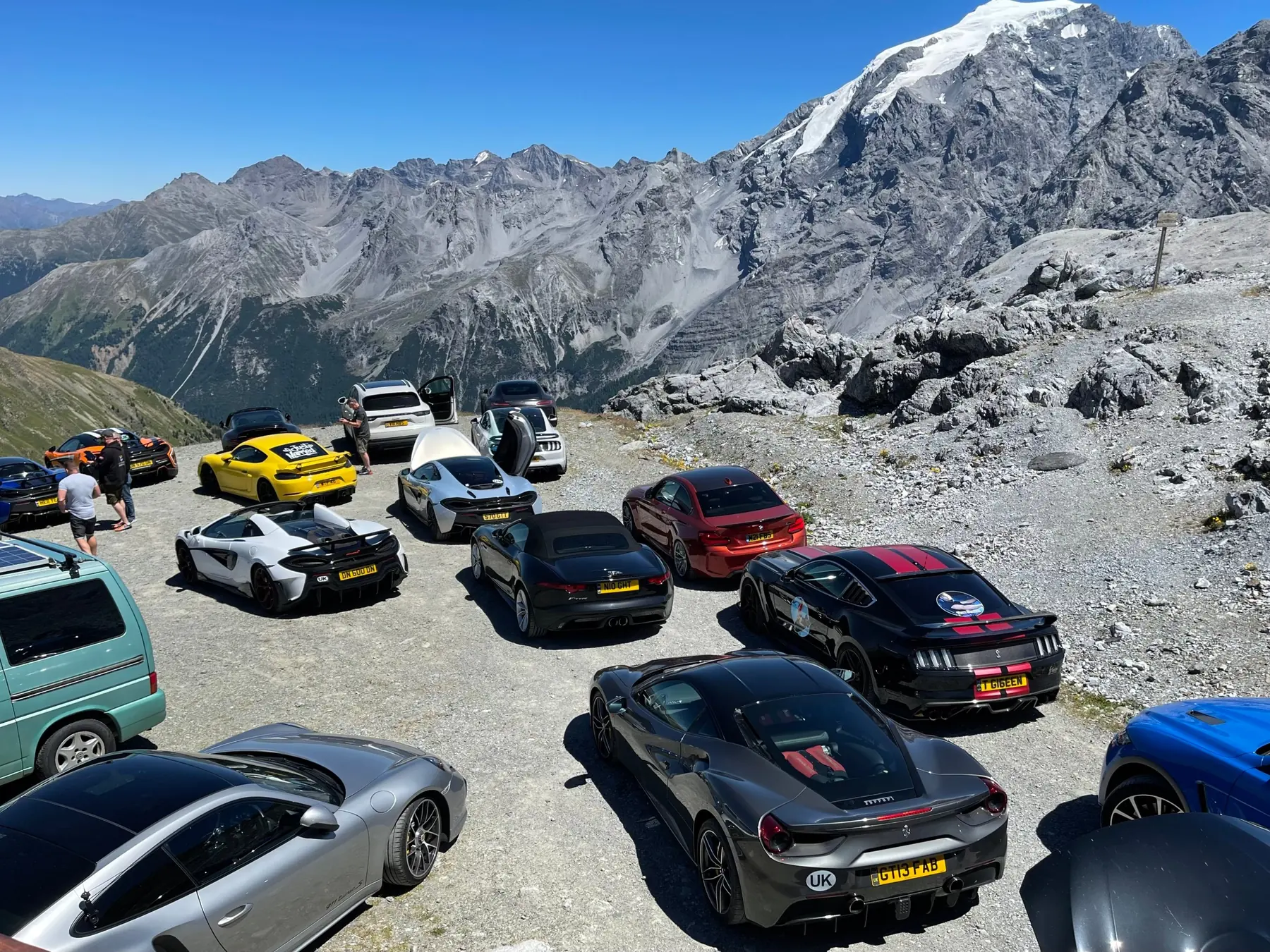 A dozen cars parked at the top of an Alpine pass in bright sun, drivers standing between them, a snow-capped peak behind.